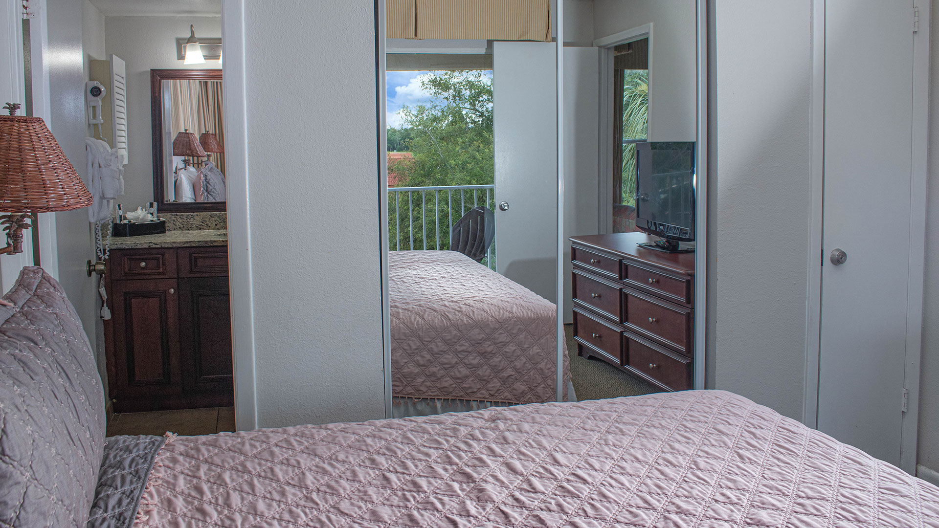 A wide view of a hotel room with a pink bedspread, a balcony, and a TV on a dresser.