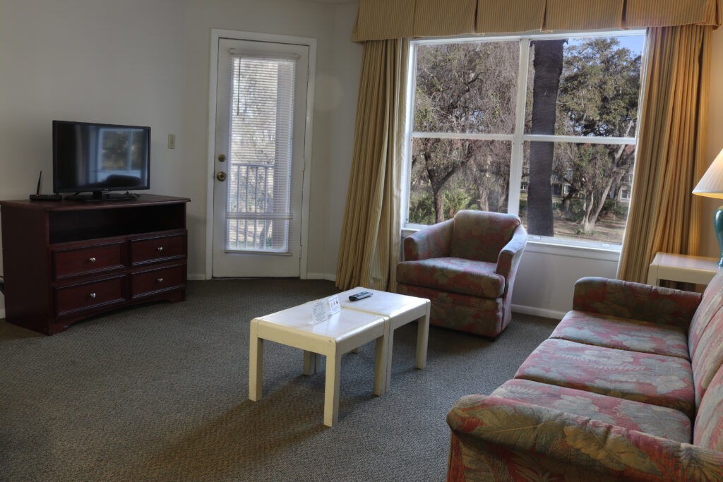 A hotel room with a floral sofa, armchair, coffee table, TV on a stand, and a window with a view.