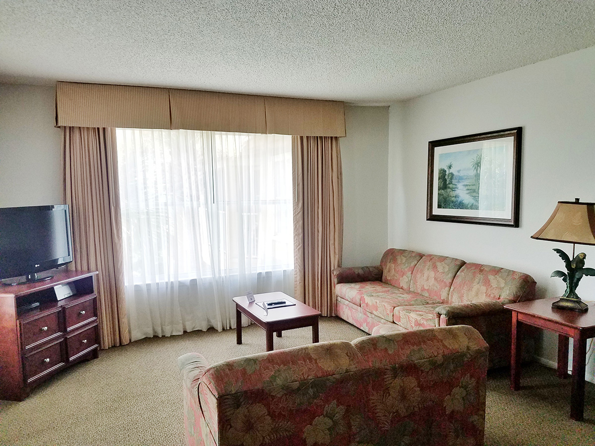 A wide view of a hotel room's living area with a sofa, armchair, coffee table, TV, and lamp.