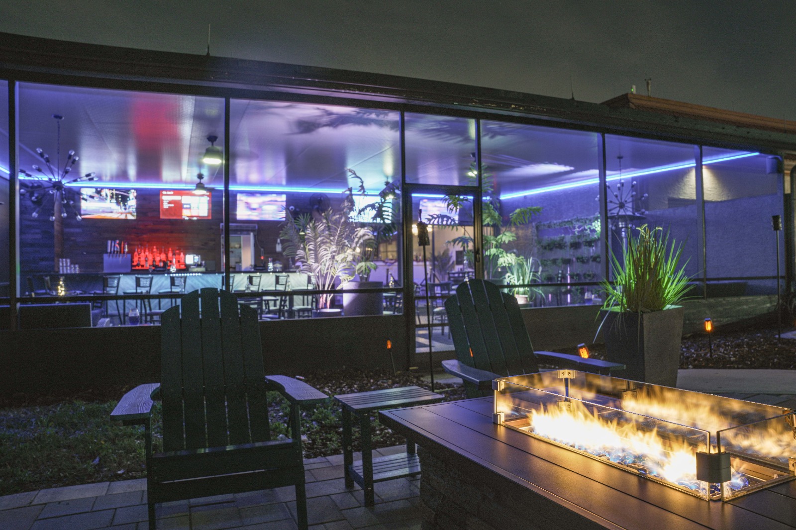 A seating area with a fire pit in front of a large windowed wall looking into a bar